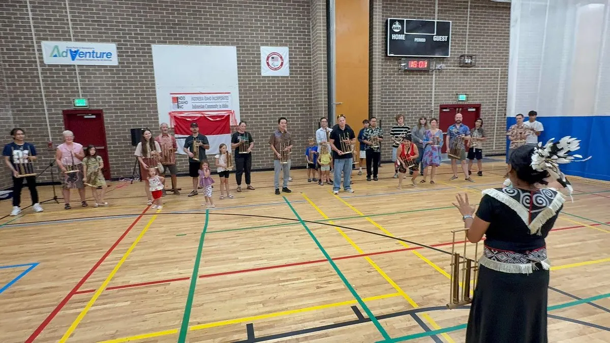Children and adults standing in a school gym holding bamboo angklung instruments while a woman in traditional dress leads the workshop